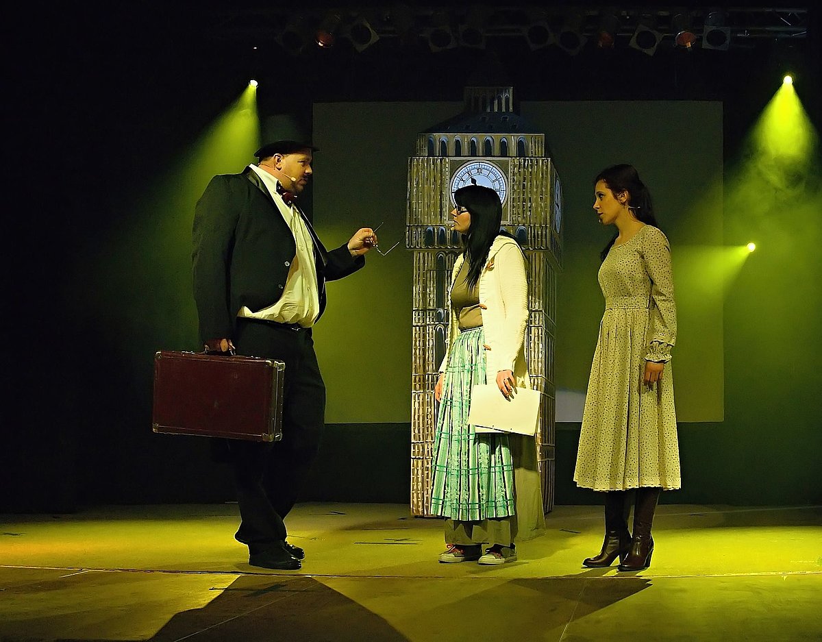 Das Foto zeigt Benjamin Baier im Anzug mit einem Koffer und zwei Frauen in Kleidern