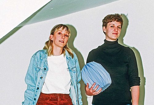 zwei Frauen stehen in einem Treppenhaus, eine hält eine Papierurne in der Hand, eine zweite Urne steht zu Füßen der Frauen