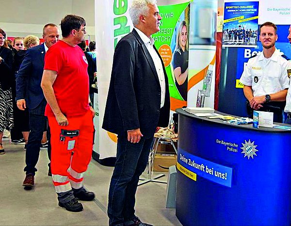 Im Vordergrund sind Bürgermeister Timo Ehrhardt mit zwei Polizeibeamten zu sehen, die sich gegenüberstehen und unterhalten. Im Hintergrund des Bildes sind mehrere Personen zu erkennen, die sich verschiedene Stände der work@ludscht anschauen.