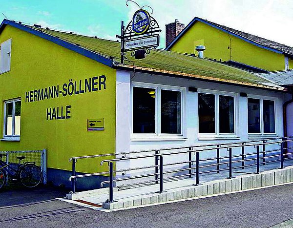 Zu sehen ist die neu gestaltete Hermann-Söllner-Halle. Die Front strahlt in einem hellen Gelb mit Schriftzug Hermann-Söllner-Halle. Rechts davon ist der barrierefreie Zugang zur Halle.