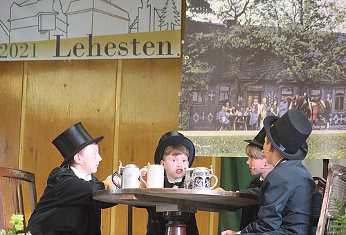 Vier Kinder in schwarzen Anzügen mit Zylinder beim Theaterstück sitzen um einen Tisch herum.
