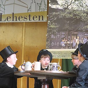 Vier Kinder in schwarzen Anzügen mit Zylinder beim Theaterstück sitzen um einen Tisch herum.
