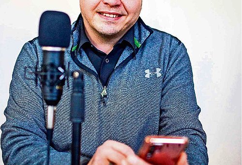 lächelnder Mann sitzt vor einem Mikrofon und hält ein Smartphone in der Hand