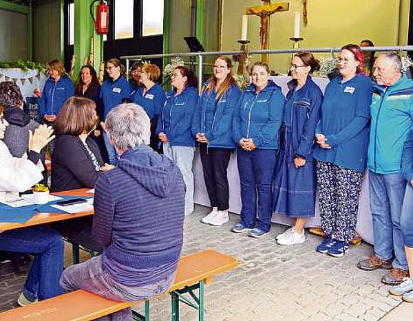 In einem Raum sitzen Menschen an Biertischgarnituren. Ganz vorn stehen Frauen in blauen Shirts in einer Reihe nebeneinander. Die Menschen klatschen für die Frauen.