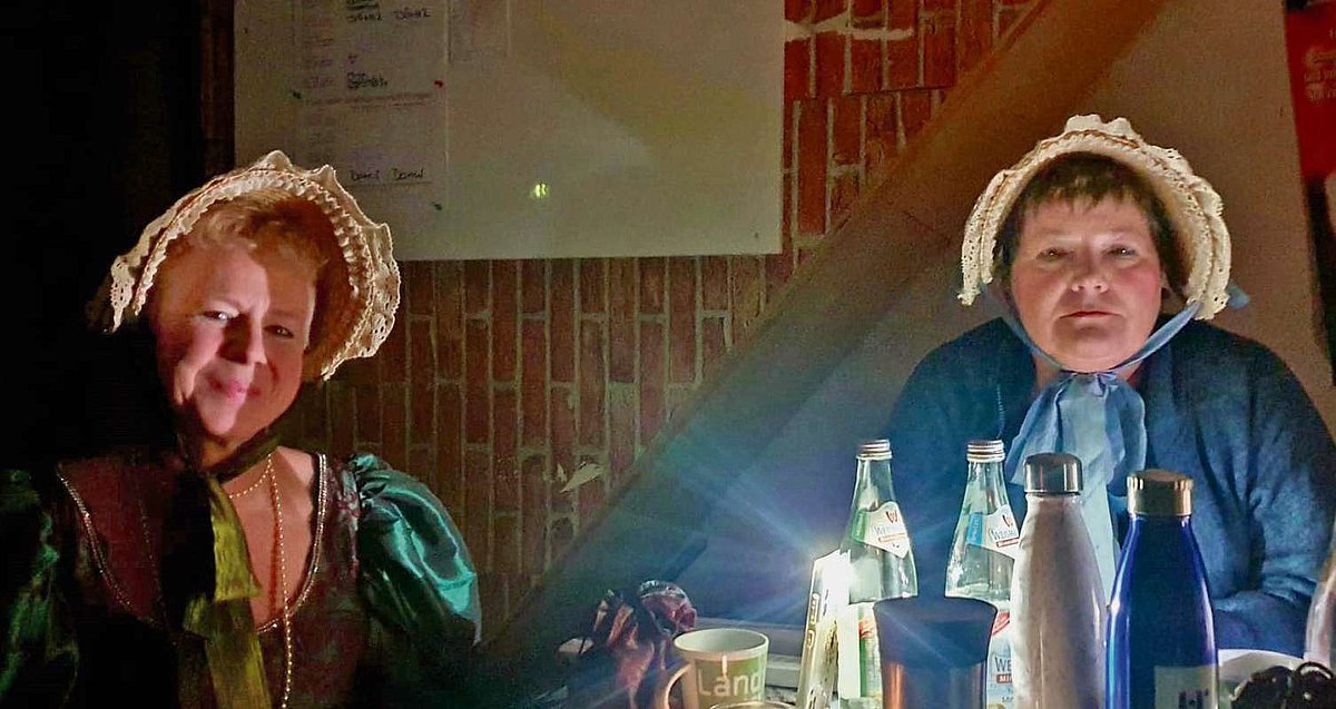 Zwei Frauen sitzen mit ihren Theaterkostümen bei Taschenlampenlicht am Tisch und schauen in die Kamera. 